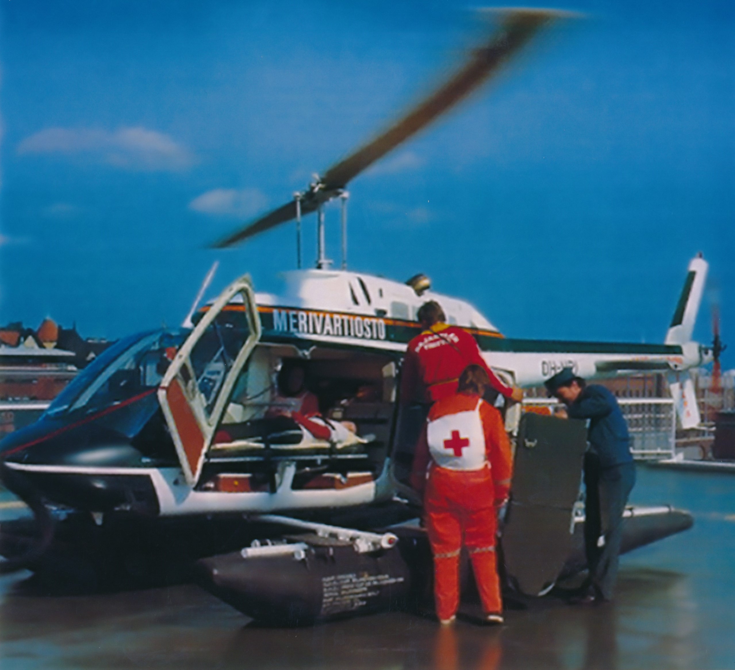 An ambulance helicopter has landed on a hospital roof. The helicopter pilot is packing up foldable stretchers. Two paramedics are standing in front of the helicopter. The patient is lying on a stretcher inside the helicopter.