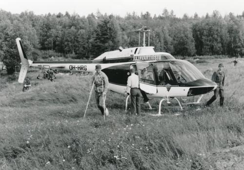 A helicopter has landed in a field. The helicopter pilot is holding the door open. Walking behind the helicopter is a uniformed soldier. Walking in front of the helicopter is an injured conscript on crutches.