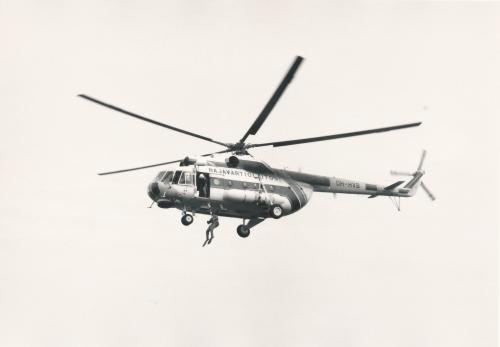 Maanpinnalta ylöspäin otettu valokuva pelastushelikopterista. Helikoperin ovella seisoo miehistön jäsen, joka käyttää vinssiä. Vinssin varassa laskeutuu pintapelastaja.