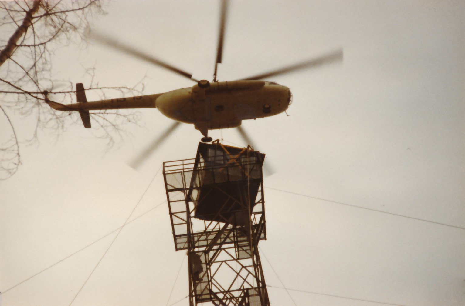 Maanpinnan tasolta ylöpäin otettu kuva helikopterista, joka pystyttää tähystystornin koppia.