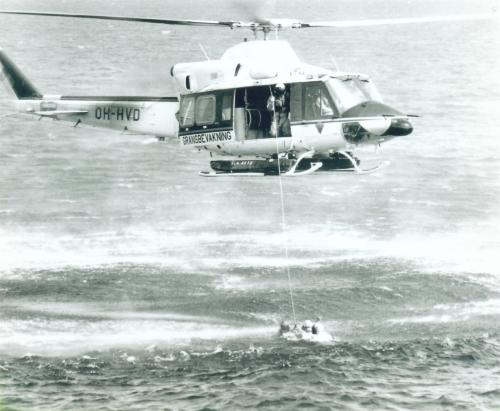 Helikopteri leijunnassa matalalla meren yllä. Kopterin oviaukko on auki ja yksi miehistön jäsen käyttää vinssiä. Vinssin varassa veteen on laskettu pintapelastaja, joka auttaa veden varassa olevaa ihmistä.