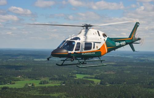 Aerial photograph of a flying helicopter. The colours of the helicopter are white, green and orange. The helicopter also has the text Rajavartiolaitos (Finnish Border Guard). 