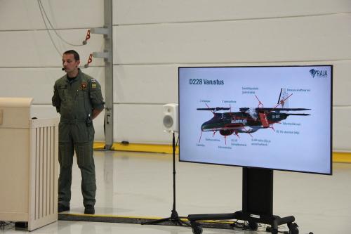 Photograph from the media briefing. On the left in the photograph, a man dressed in flying gear is speaking to the audience. On the right, there is a large television screen showing a diagram of the aeroplane.