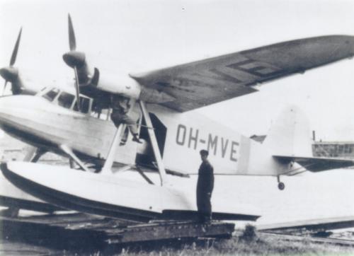 A twin-engine aeroplane with floats that has been brought onto land for maintenance work. A man is standing in front of the aeroplane. The mechanic is servicing one of the aeroplane’s engines.