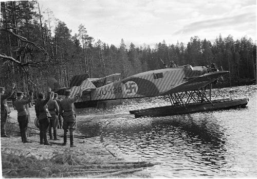 A photograph of a seaplane on the shore of a lake during the Continuation War. The aeroplane is being prepared for take off. On the shore, a group of people are standing and waving to the aeroplane.