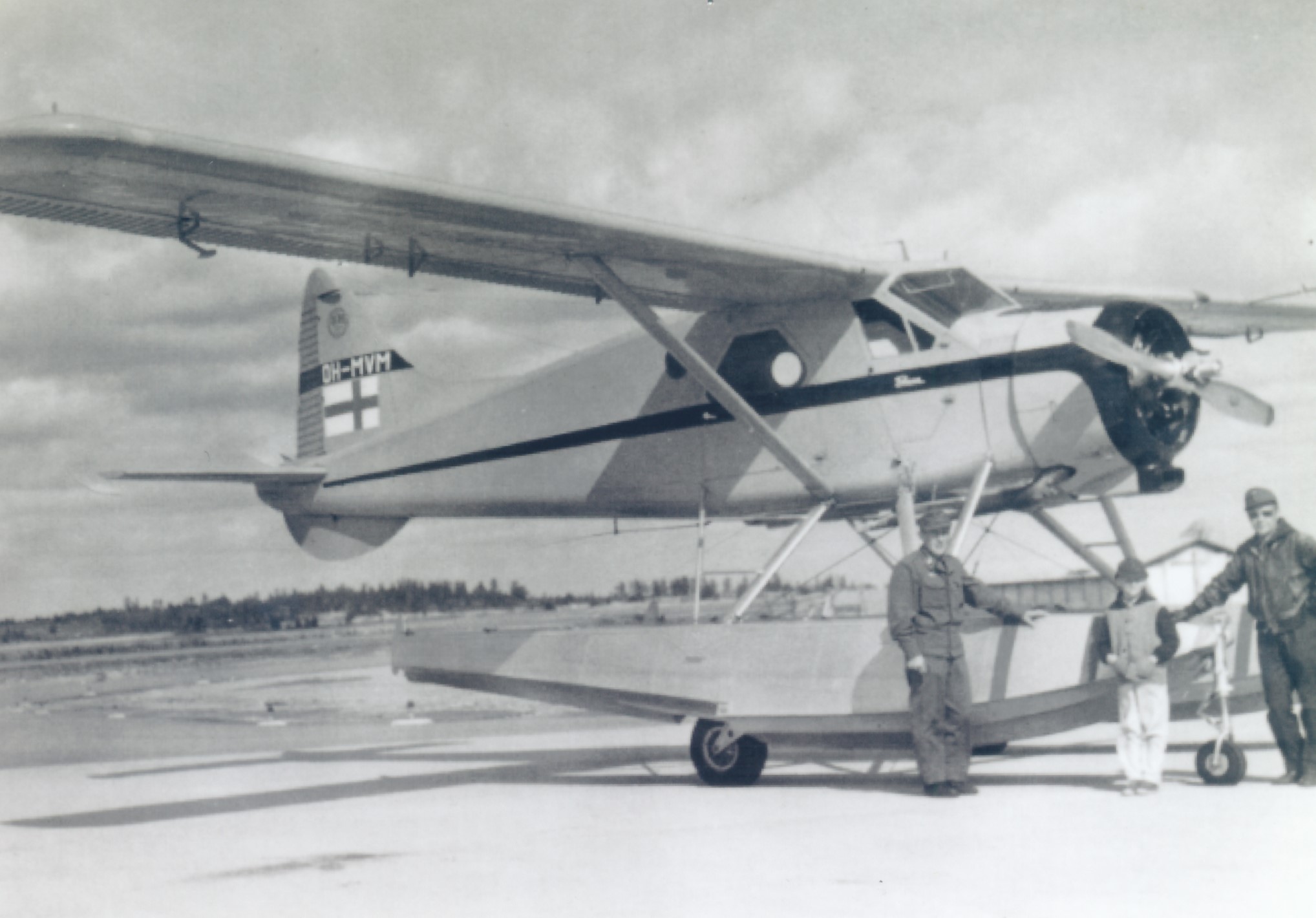 A single-engine aeroplane with floats stands on the asphalt of an airstrip. Two crew members are standing in front of the plane and a small boy is standing between them.