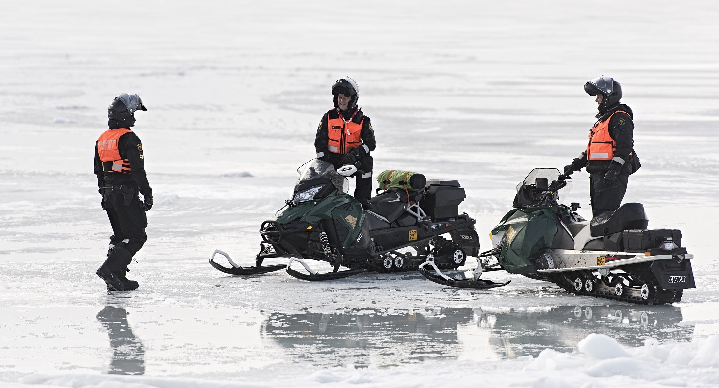 Kolme miestä huomioliivit päällä moottorikelkkojen vieressä jäällä.