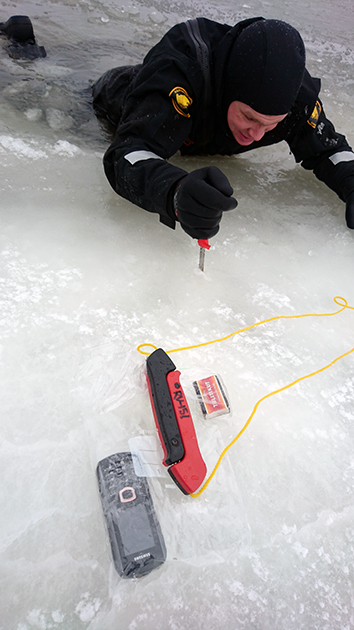 En sjöbevakare tar sig upp ur vaken med hjälp av isdubbar. En påse packad med mobiltelefon och tändsticksask på isen.