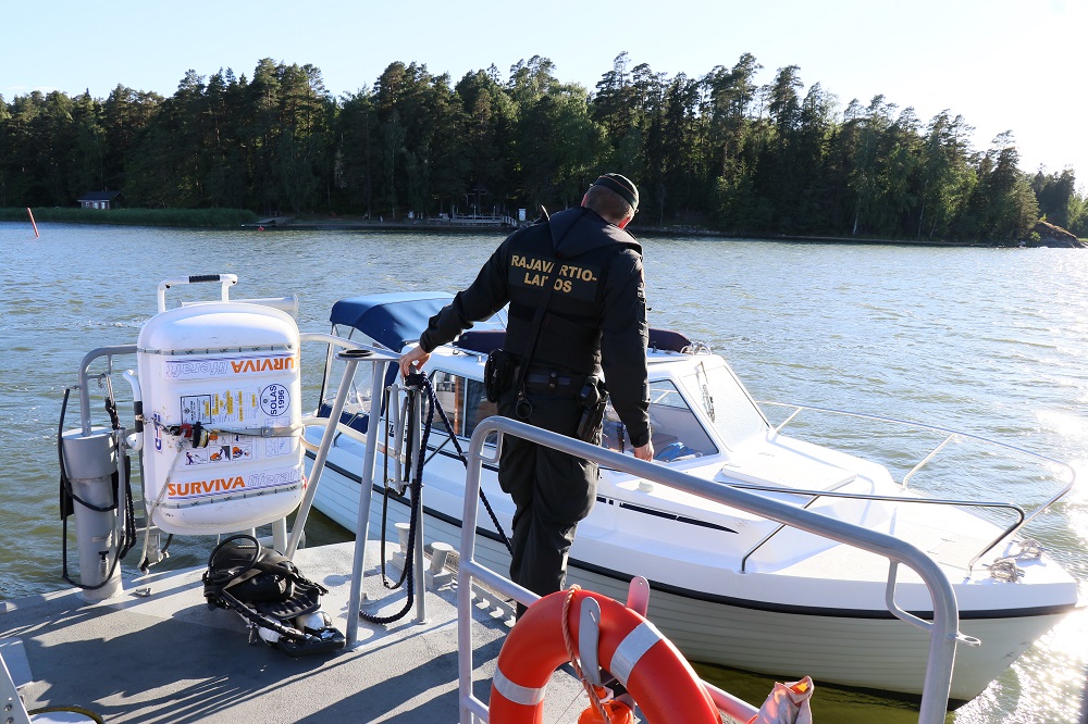 Rajavartija seisoo partioveneen takakannella. Partioveneen vieressä huvivene.