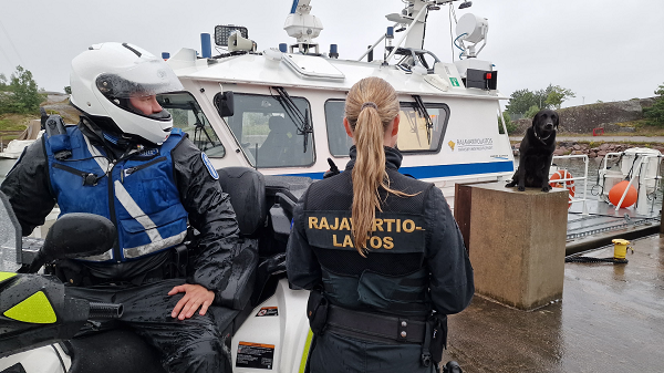 Partioveneen edessä laiturilla poliisin virkamies mönkijän kyydissä ja selin kameraan Rajavartiolaitoksen virkamies. Katsovat Rajavartiolaitoksen koiraa, joka on puulaatikon päällä.