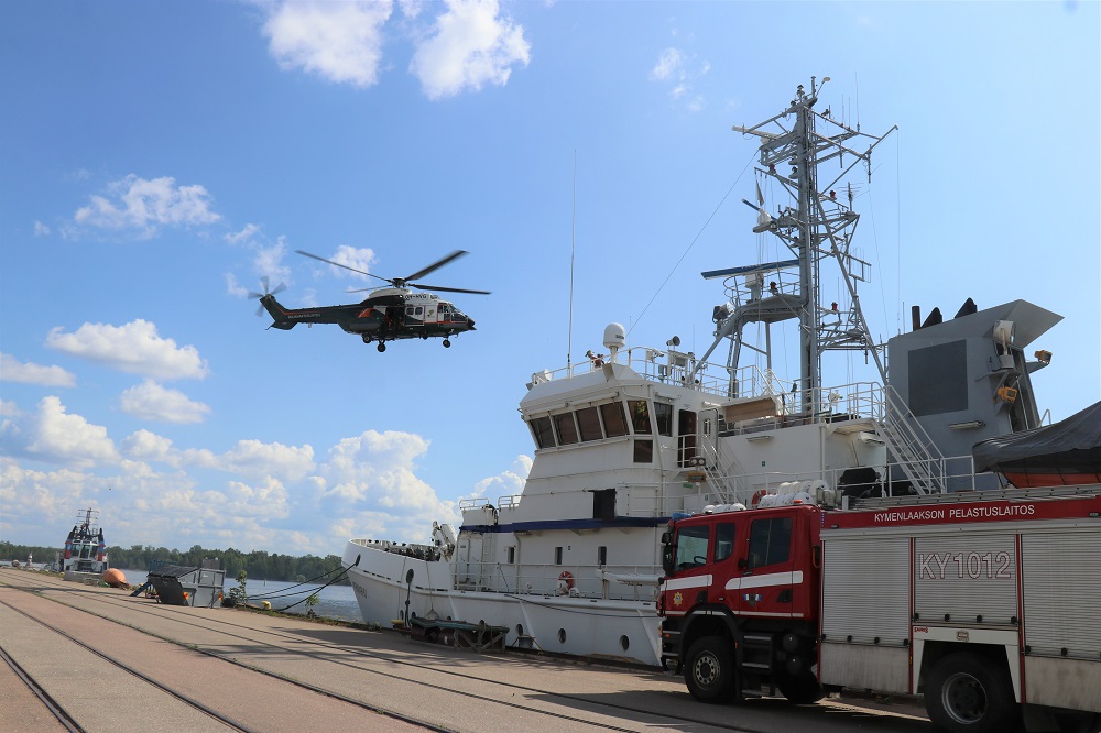 Helikopteri leijumassa laivan yläpuolella. Laiva on satamassa kiinni ja vieressä on paloauto.