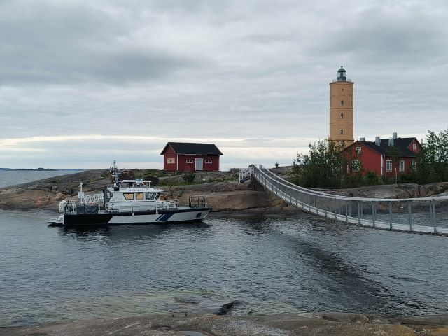 Partiovene saaren laiturissa. Saarelle johtaa silta ja, siellä on muutama rakennus.