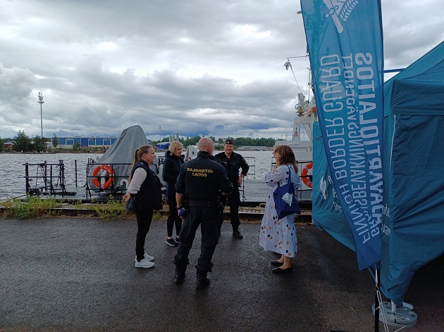 Kolme henkilöä keskustelee merivartijoiden kanssa teltan edessä satamassa.