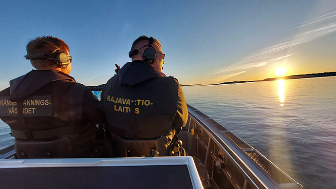 Kaksi virkamiestä partioveneessä merellä ja auringonlasku oikeassa reunassa.