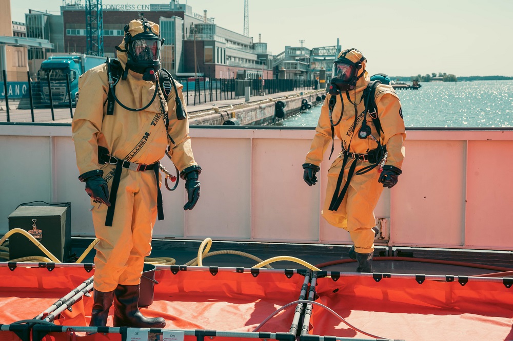 Kaksi henkilöä kemikaalipuvut päällä laivan kannella.