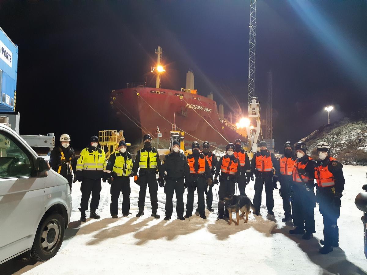 Laiva taustalla, edessä rajan ja tullin virkamiehiä huomiliivit päällä, yksi merivartiokoira