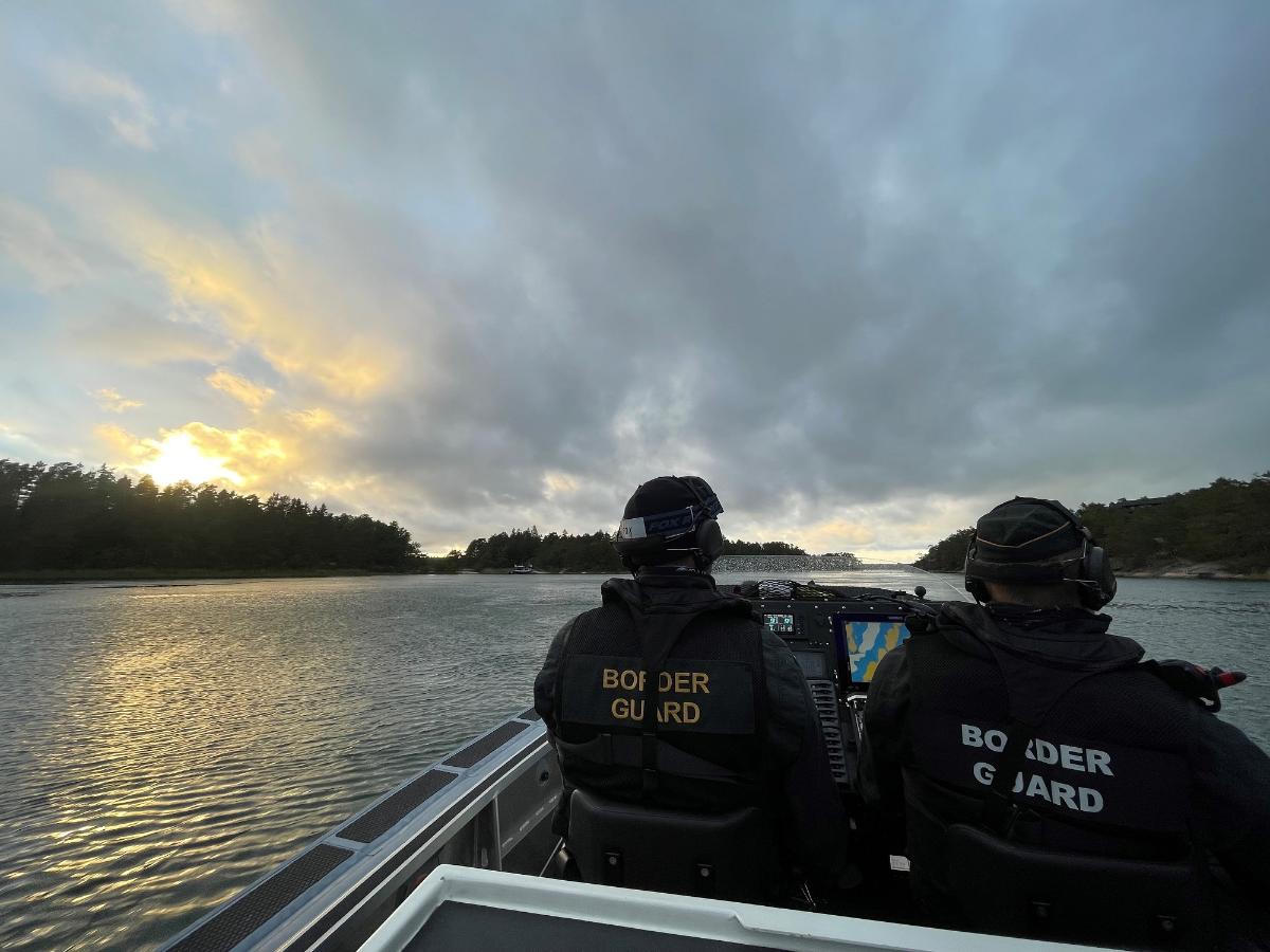 Kaksi merivartioston virkamiestä vierekkäin partioveneessä. Kuva otettu takaapäin ja selässä näkyy , henkilöt katsovat eteenpäin. Meri tyyni.