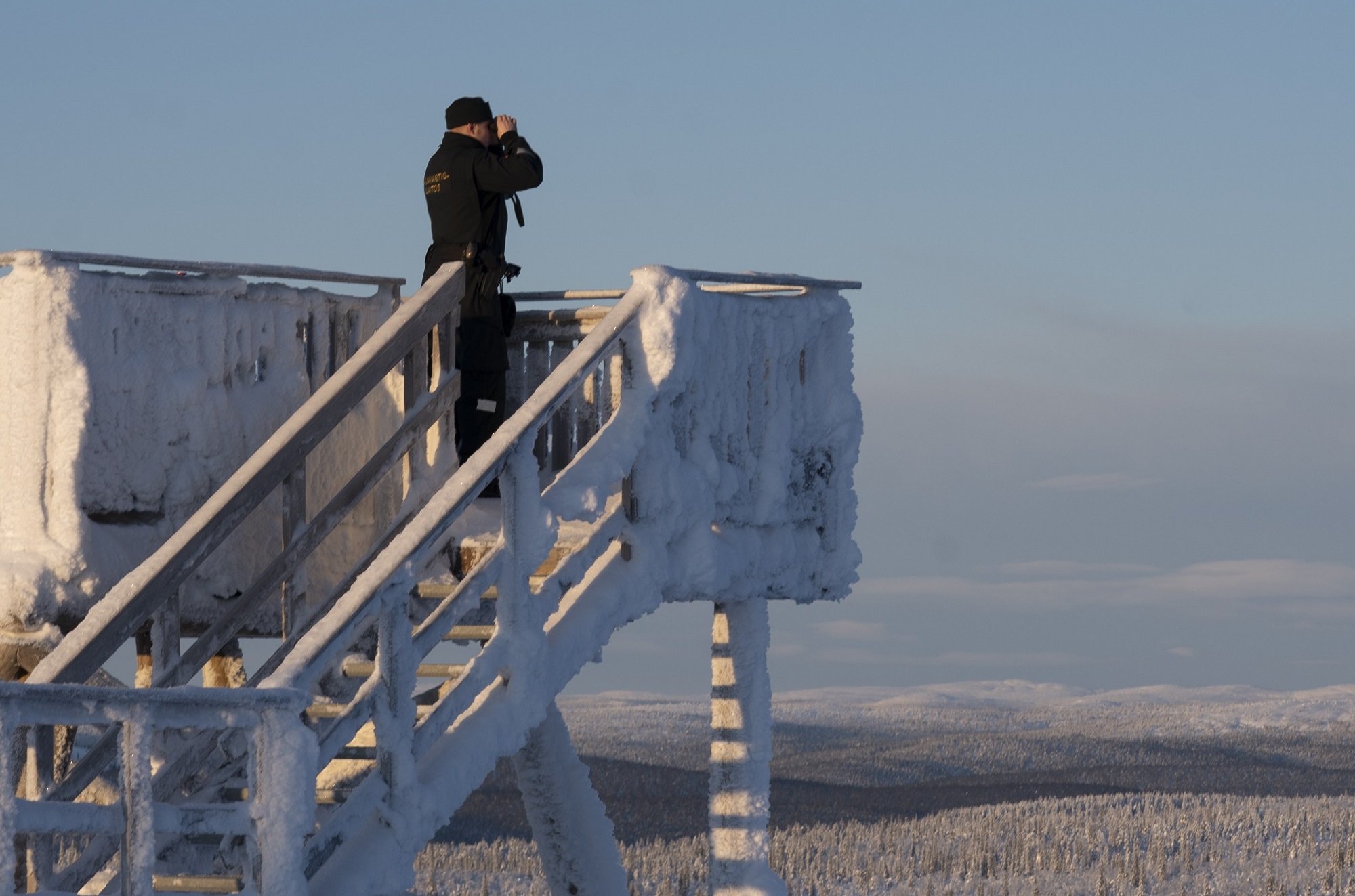 A border guard with binoculars looking out from a snowy observation tower.