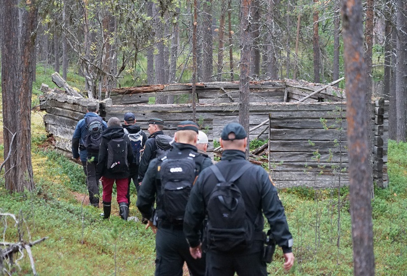 Seitsemän ihmistä kulkee jonossa selkä kameraan päin kohti metsässä sijaitsevaa kämpän rauniota.