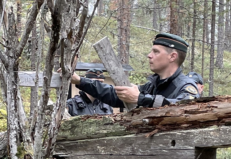 Upseeri Jarkko Alén tutkii kämpän oven päällä aikoinaan ollutta kylttiä, joka on ripustettuna puuhun.