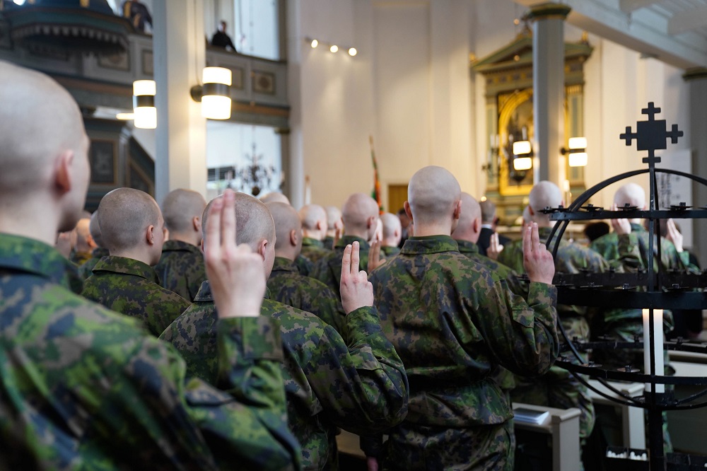 Varusmiehiä seisoo kirkossa oikean käden kaksi sormea ylhäällä. Kuva otettu takaapäin.