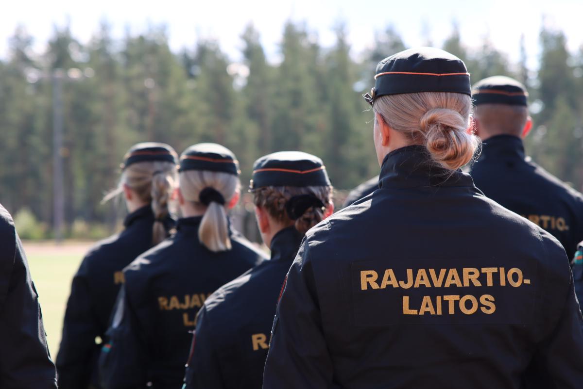 Rajavartijaopiskelijoita seisomassa. Kuva otettu takaapäin. Takeissa lukee 