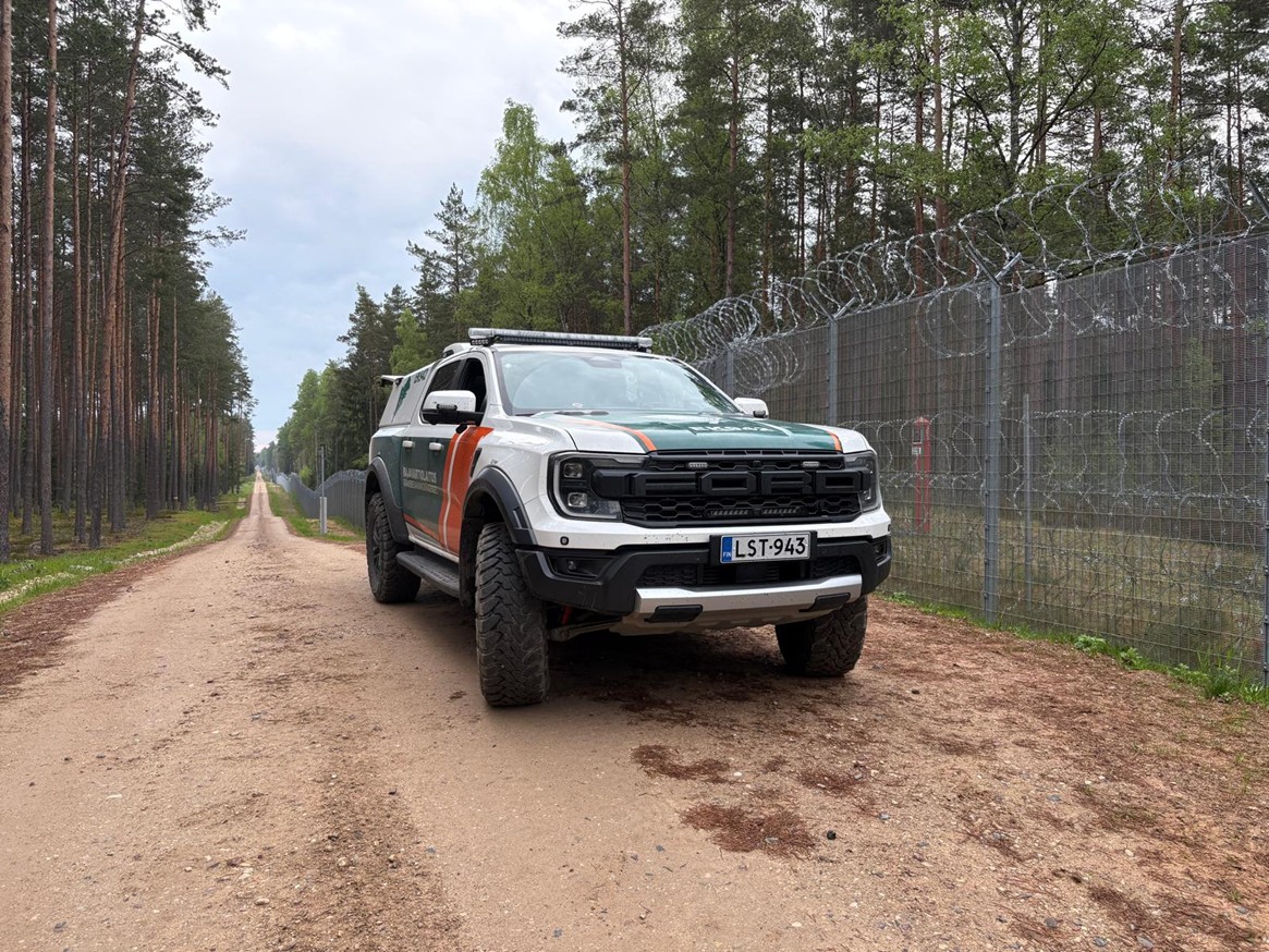 Suomen Rajavartiolaitoksen auto seisoo Latvian rajalla hiekkapäällysteisellä tiellä. Taustalla on metallinen verkkoaita ja metsää.