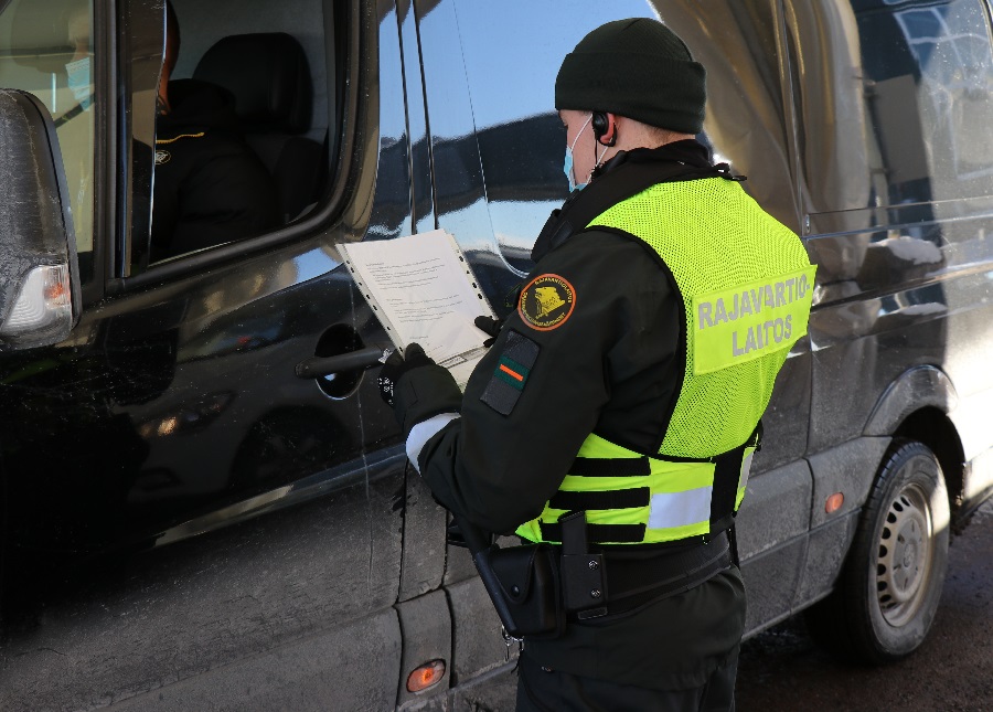Gränsbevakare i tjänsteuniform och skyddsmask granskar resedokument utanför en svart bil och en person med skyddsmask sitter i bilen i chaufförssätet.