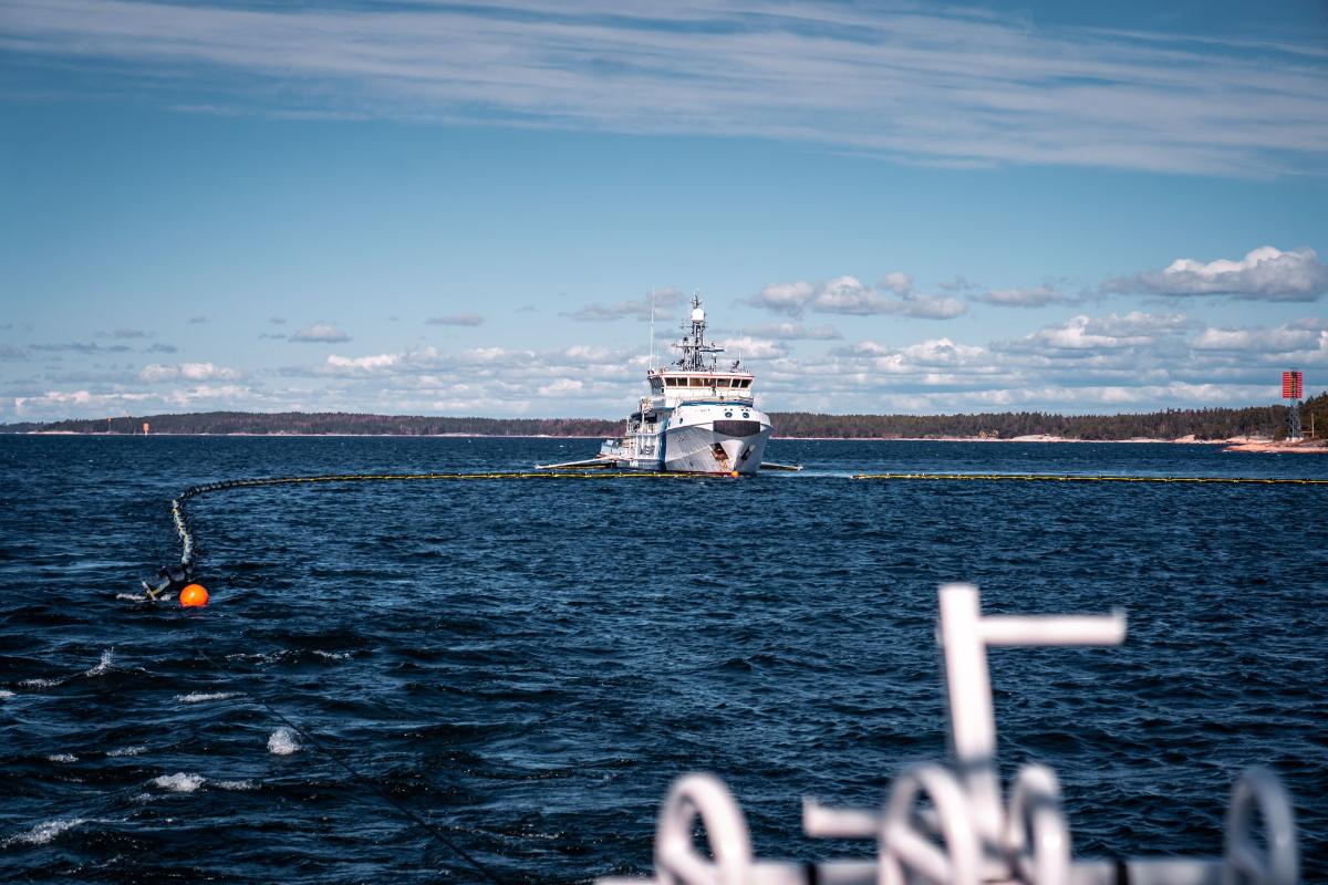 Booms are being towed at sea. In the background there is one of the offshore patrol vessels of the Finnish Border Guard.